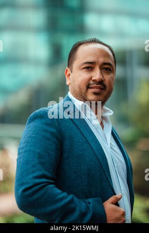 Side view of Hispanic mature male manager in suit holding hands in pockets and looking at camera with smile while walking near glass building on sunny Stock Photo