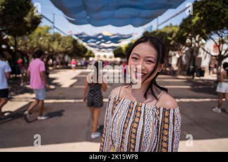 Cheerful young Asian female smiling and looking at camera while standing on blurred background of crowded park on sunny day in Seville, Spain Stock Photo