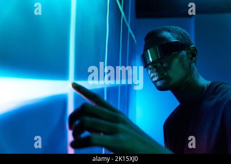 Concentrated young African American male in contemporary VR glasses touching large projector screen with geometric pattern while exploring cyberspace Stock Photo