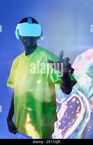 Concentrated young African American in green t shirt and modern VR goggles pointing and touching screen while interacting with virtual object in dark Stock Photo