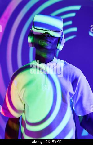 Stylish young African American male millennial in white t shirt experiencing virtual reality in modern VR headset in studio with colorful neon illumin Stock Photo