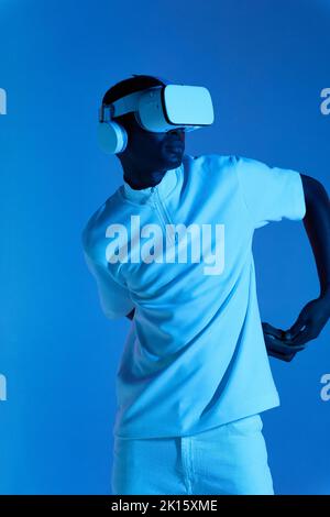 Cool young African American male in white outfit and futuristic VR goggles with headphones looking away while standing against blue background with ha Stock Photo