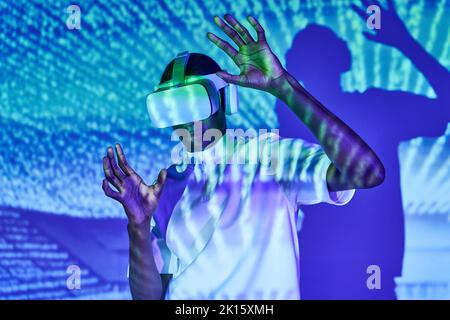 Focused young African American in white t shirt touching imaginary object while exploring virtual reality in modern headset against wall with bright p Stock Photo