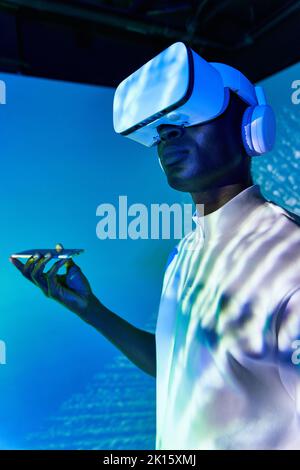 Confident young African American male in casual clothes and VR goggles using smartphone while standing in studio with bright neon illumination Stock Photo