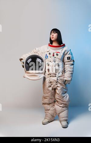 Young Asian woman in spacesuit holding helmet and looking away under blue light before mission against gray background Stock Photo
