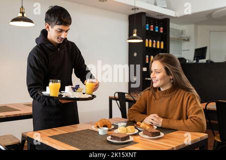 Young man in black uniform taking glass of juice from tray while serving food for smiling woman during breakfast in restaurant Stock Photo