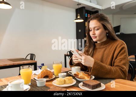 Young lady in warm sweater smiling and messaging on mobile phone while sitting at table with toasts and beverages during breakfast in cafe Stock Photo