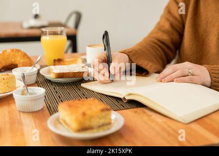 Cropped unrecognizable woman in brown sweater sitting at table with breakfast food and drinks and writing in planner while working remotely during bre Stock Photo