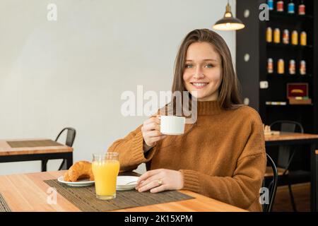 Cheerful young female in sweater enjoying coffee and looking at camera with smile while sitting at table with croissants and juice in light restaurant Stock Photo