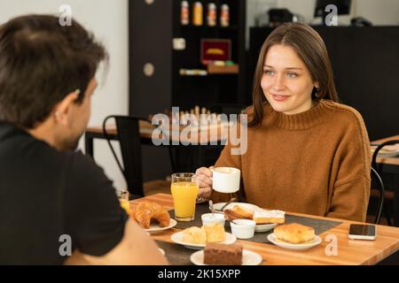 Happy young woman enjoying coffee and listening to boyfriend while having breakfast in restaurant together Stock Photo