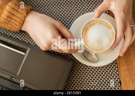 Top view of anonymous female freelancer taking cup of latte from saucer near laptop during coffee break in cafe Stock Photo