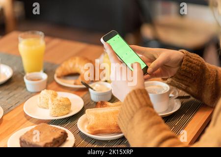Cropped unrecognizable female in casual clothes using smartphone with chromakey screen to take photo of assorted breakfast food and drinks in restaura Stock Photo
