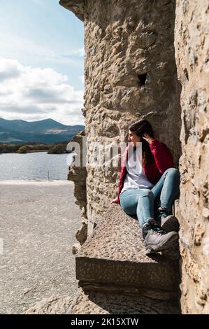 Full body woman in casual clothes sitting on stone bench near aged building and looking away on sunny day on shore Stock Photo