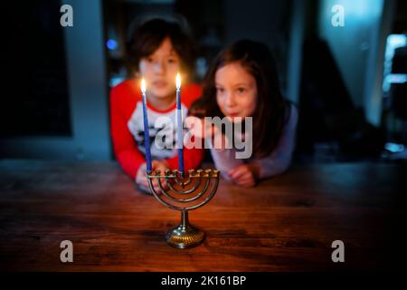 Two siblings sit at table together staring at lit candles on menorah Stock Photo