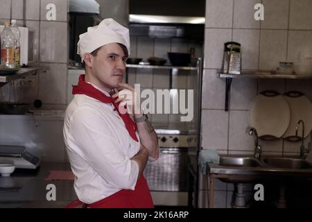 Young handsome chef posing in kitchen in restaurant. Portrait of positive male cook in uniform Stock Photo