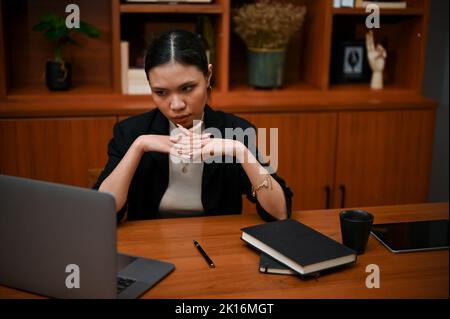 Thoughtful and determined young Asian businesswoman or female boss looking at laptop screen, focused on planning her business strategy in the office Stock Photo