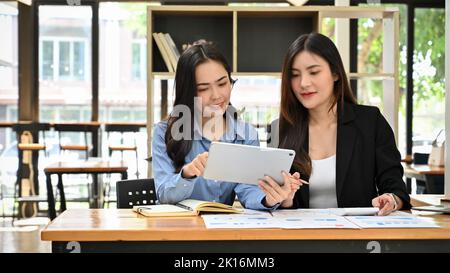Two beautiful millennial Asian businesswomen or female marketing employees remote working at the cafe, working and planning their marketing project. Stock Photo