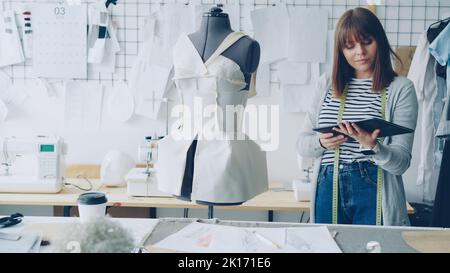 Young female fashion designing entrepreneur is working with tablet while checking clothing pinned to tailoring dummy. Modern technologies in clothes manufacturing concept. Stock Photo