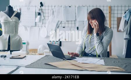 Confident clothing designer is discussing garment sketches on mobile phone and entering information in laptop. Busy day in clothes designing studio concept. Stock Photo