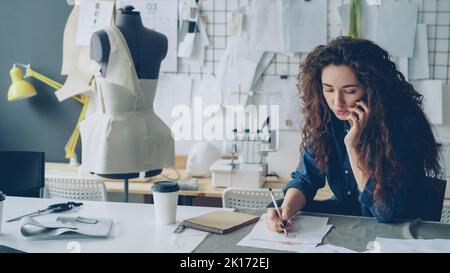 Professional female fashion designer is talking on mobile phone and drawing clothing sketch with pencil at table in tailoring shop. Everyday work and communication in workplace concept. Stock Photo
