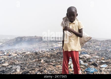 Neglected, malnourished African boy standing desolate in the midst of a smelly and smoking garbage dump; social issue of child abandon Stock Photo