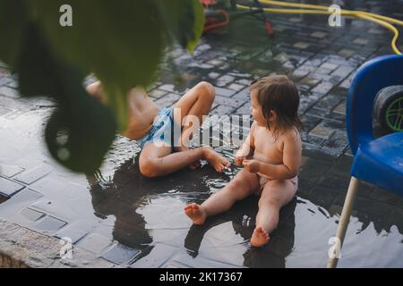 Two wet children playing with garden sprinkler on hot sunny day. Summer outdoor fun activity. Beautiful sunset. Summer vacation. Happy family. Stock Photo