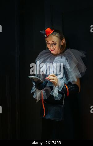 Elite clown. Smiling girl dressed as clown is browsing social networks on her phone at party. Contrast image on black background. High quality photo Stock Photo