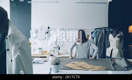 Young female designer standing with her back to camera and looking at drawings hanging on white wall. Attractive woman is thinking about new clothing collection. Stock Photo