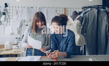 Creative dressmakers are using smartphone and looking at clothing sketches while working in nice modern studio. Dummy, sewing machine and women's garments in background. Stock Photo