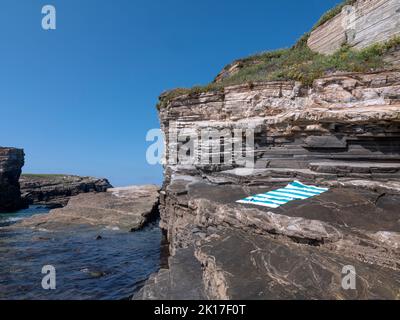 lonely striped blue towel in a rocky beach full of seaweed and cliffs in the background. Stock Photo