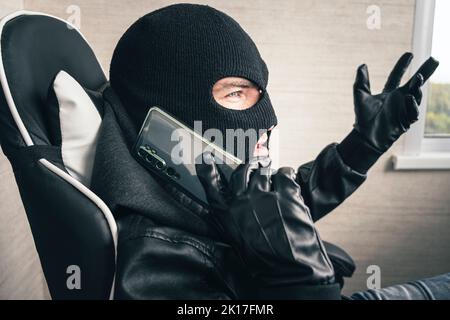 Young adult in black clothes with hidden face. Ill-intended fraudster uses mobile. Fraudster calls. Mobile racket. Hacker hijacks by phone. Cellphone Stock Photo