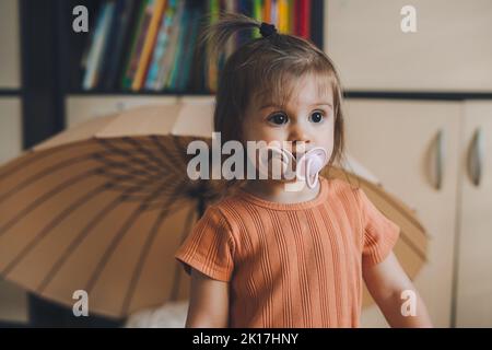 Funny portrait of a baby girl standing in the house with two pacifiers in her mouth looking to one side. Happy infant. Family people indoor Interior Stock Photo