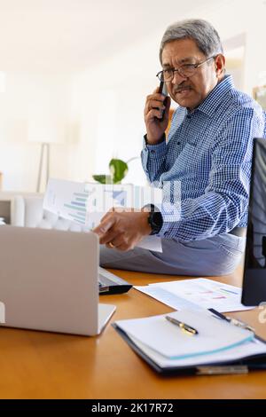 Senior biracial businessman talking on smartphone and using laptop sitting on desk at home Stock Photo