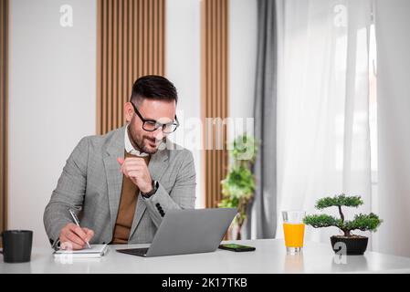 Delighted adult businessman, checking the company income on his laptop, writing it down. Stock Photo
