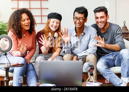 Group of positive multiracial friends waving hands while having video call via laptop on table with microphone and ring lamp Stock Photo