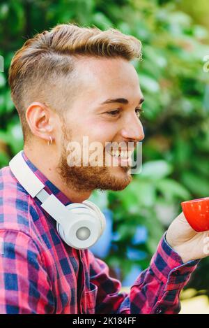 Side view of positive bearded male with modern wireless headphones drinking hot espresso while sitting on terrace of outdoor cafeteria Stock Photo