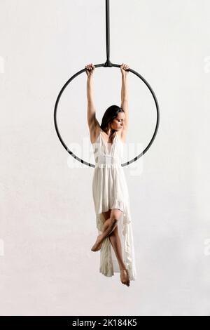 Full length of strong gracious female performer in white dress hanging from aerial hoop and looking away during rehearsal in light studio Stock Photo