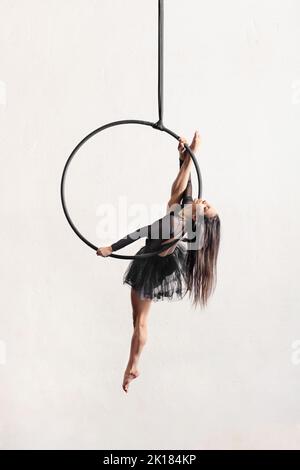 Full body barefoot dancer with long dark hair doing split pose on aerial ring during performance against gray background Stock Photo