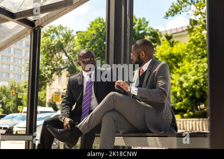 Two Two dark-skinned businessmen in suits communicate. Friends and business partners are sitting on a bench in the city against the backdrop of Stock Photo