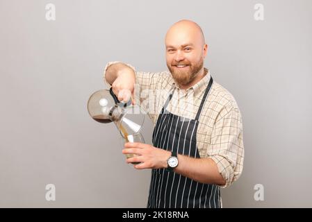 Happy bald barista man is pouring come coffee from chemex coffeemaker over light grey background. Stock Photo