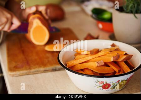 Enamel vintage bowl with sweet potato wedges against blurred chef cutting whole tubers of organic batata on wooden board Stock Photo