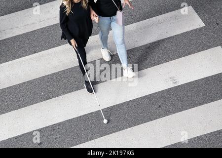 Scene of a Blind woman walking on zebra crossing helped by another person using her white cane. Help in the early stages of blindness Stock Photo