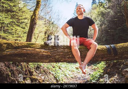 Smiling middle-aged man sitting on the fallen tree log over the mountain forest stream with his beagle dog while he waiting for laundry drying and tre Stock Photo