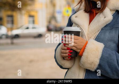 Paper cup with coffee in hands of unrecognizable woman, standing in city street. Body part of female in warm clothes with bright manicure holding hot Stock Photo