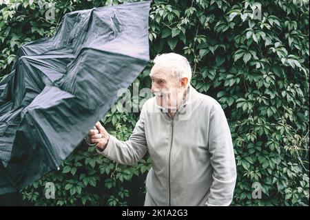 An old 88-year-old man with an umbrella on a background of green foliage. Portrait of an elderly man who opens a large black umbrella. Stock Photo