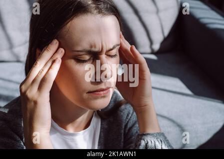 Close up of stressed young caucasian woman touching head and feels dizzy, suffering from headache and migraine pain, sitting on grey sofa at home, tir Stock Photo