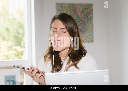 Young female remote employee in white bathrobe with earphones sitting at table with laptop and communicating with client via smartphone while working Stock Photo