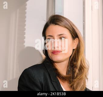 Positive young female with long brown hair wearing formal black blazer standing near window Stock Photo
