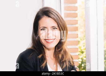 Cheerful attractive young businesswoman in formal clothes looking at camera and smiling friendly while standing near window in light room Stock Photo