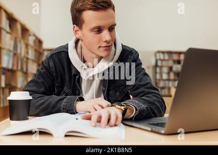 Male student sitting at desk looking at laptop. Young guy in library preparing exams. Stock Photo